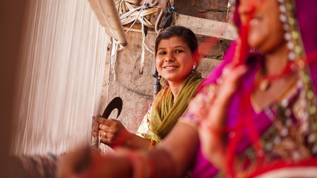 A young woman works at a loom and looks at the camera to smile.