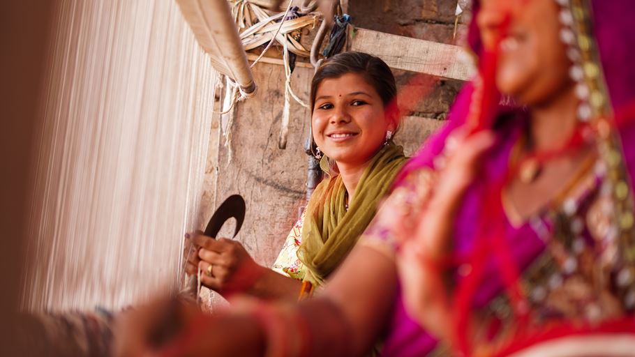A young woman works at a loom and looks at the camera to smile.