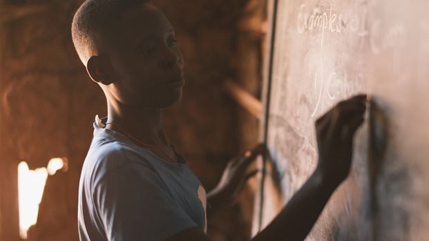 A young student writes examples on a chalkboard in a classroom, focused and determined.