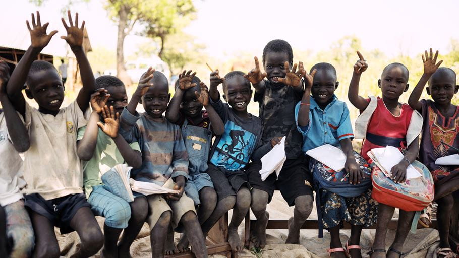 A group of South Sudanese children smile and wave at the camera, some holding school supplies.