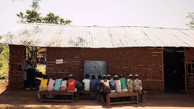 Children sit on benches facing a blackboard at an outdoor mud brick school.