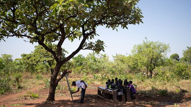 Outdoor classroom setting with children learning under a tree, instructor using a chalkboard.