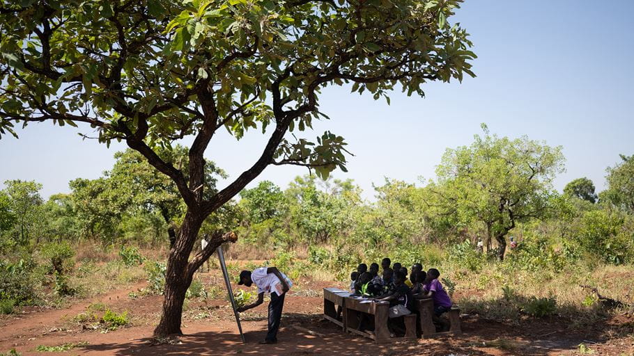 Outdoor classroom setting with children learning under a tree, instructor using a chalkboard.