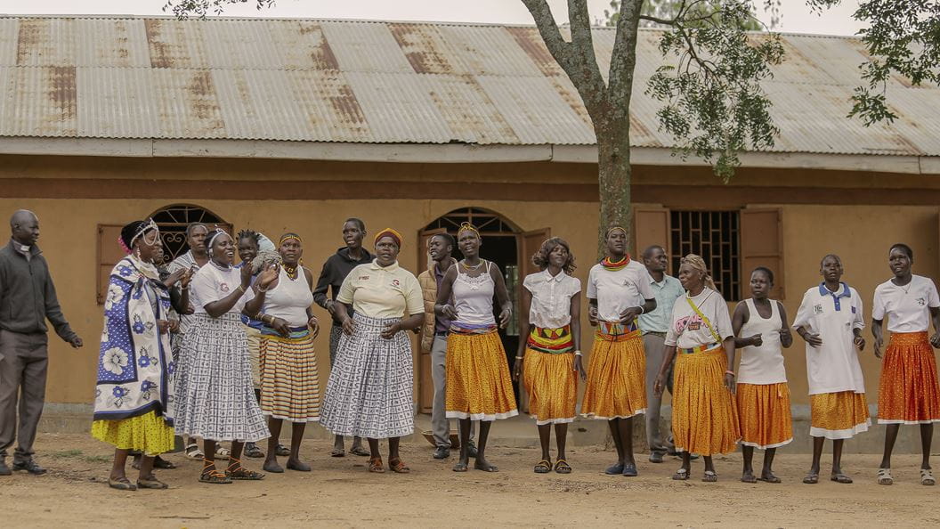A group of people dressed in long patterned skirts sing and dance in front of a building. To the left of them is a man wearing a clerical collar.