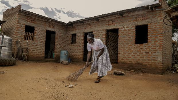 A woman wearing a white tshirt and a long skirt sweeps the ground outside a brick building with small tree branches. 