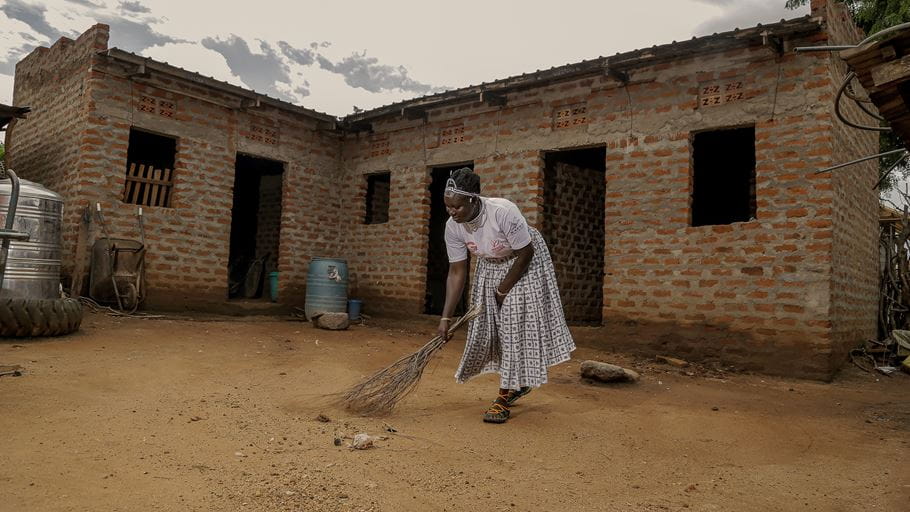 A woman wearing a white tshirt and a long skirt sweeps the ground outside a brick building with small tree branches. 