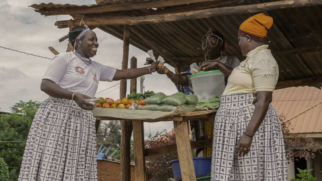 A woman wearing a white tshirt and long skirt smiles as she looks at a woman stood at a wooden stool with vegetables. Another woman behind the stool passes her a small bag.