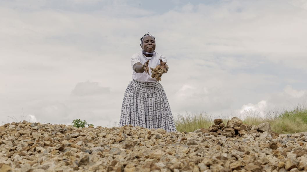 A woman wearing a white tshirt and long skirt throws rocks onto a large pile of rocks in the foreground.