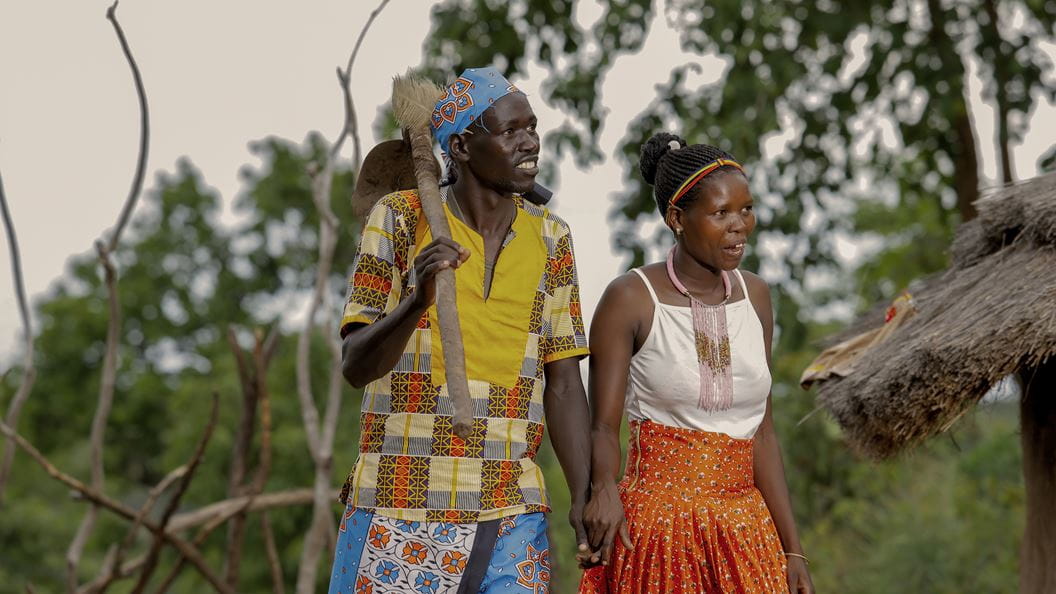 A man and woman hold hands, the man has a tool slung over his shoulder. Behind them out of focus are some trees and the corner of a straw roof. 