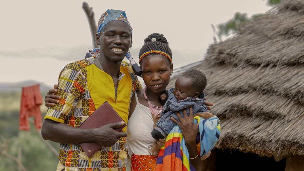 A man puts his arm around a woman, who is holding a child in her arm. The man holds a bible with his other arm. In the background, out of focus is a straw roof.  