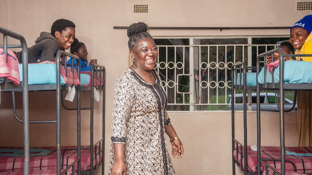 A woman who is smiling is stood between several bunkbeds either side of her. Each bunkbed has a child lying on the top and smiling.