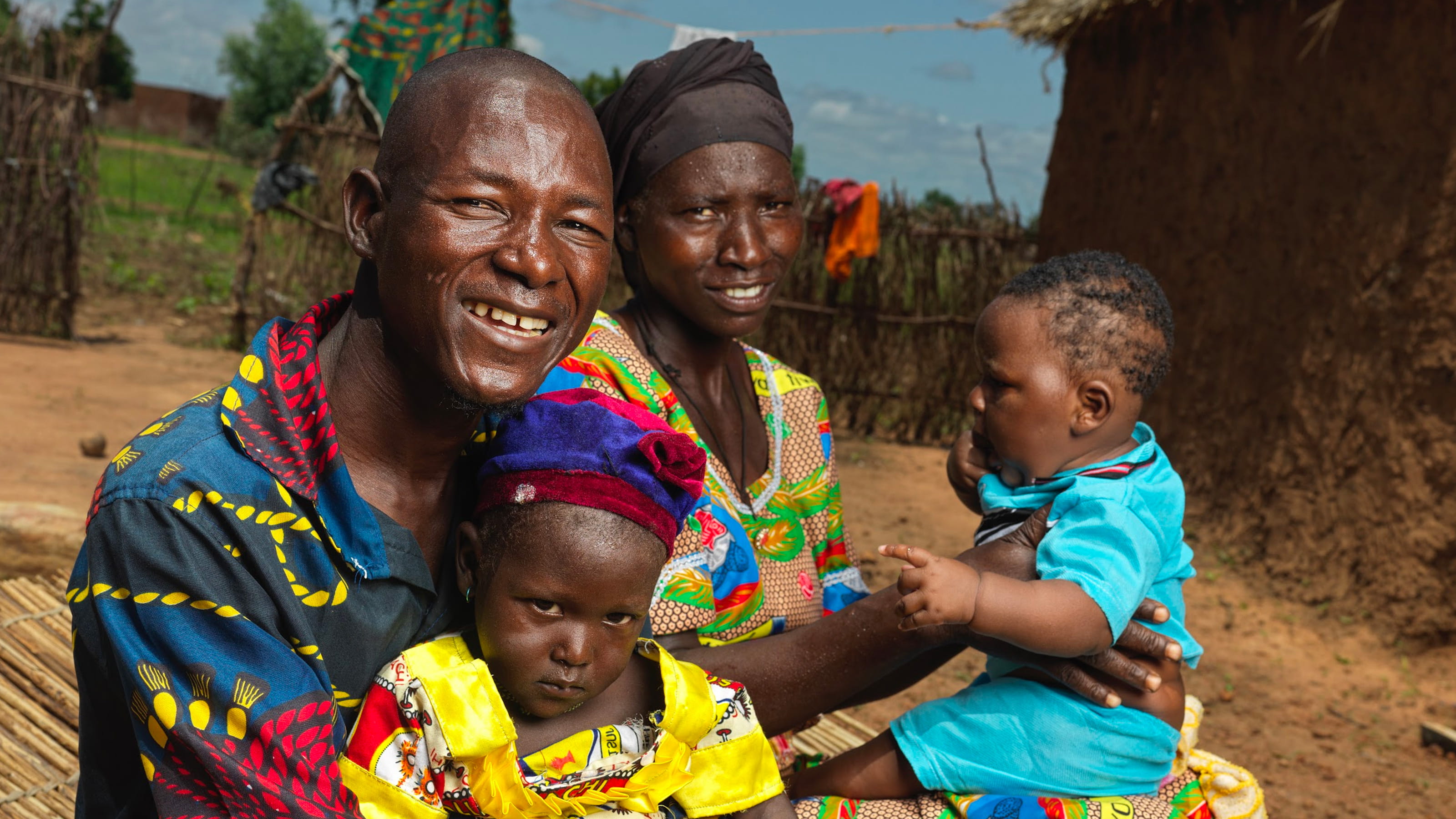 A man and woman are smiling and holding two small children outside a hut.