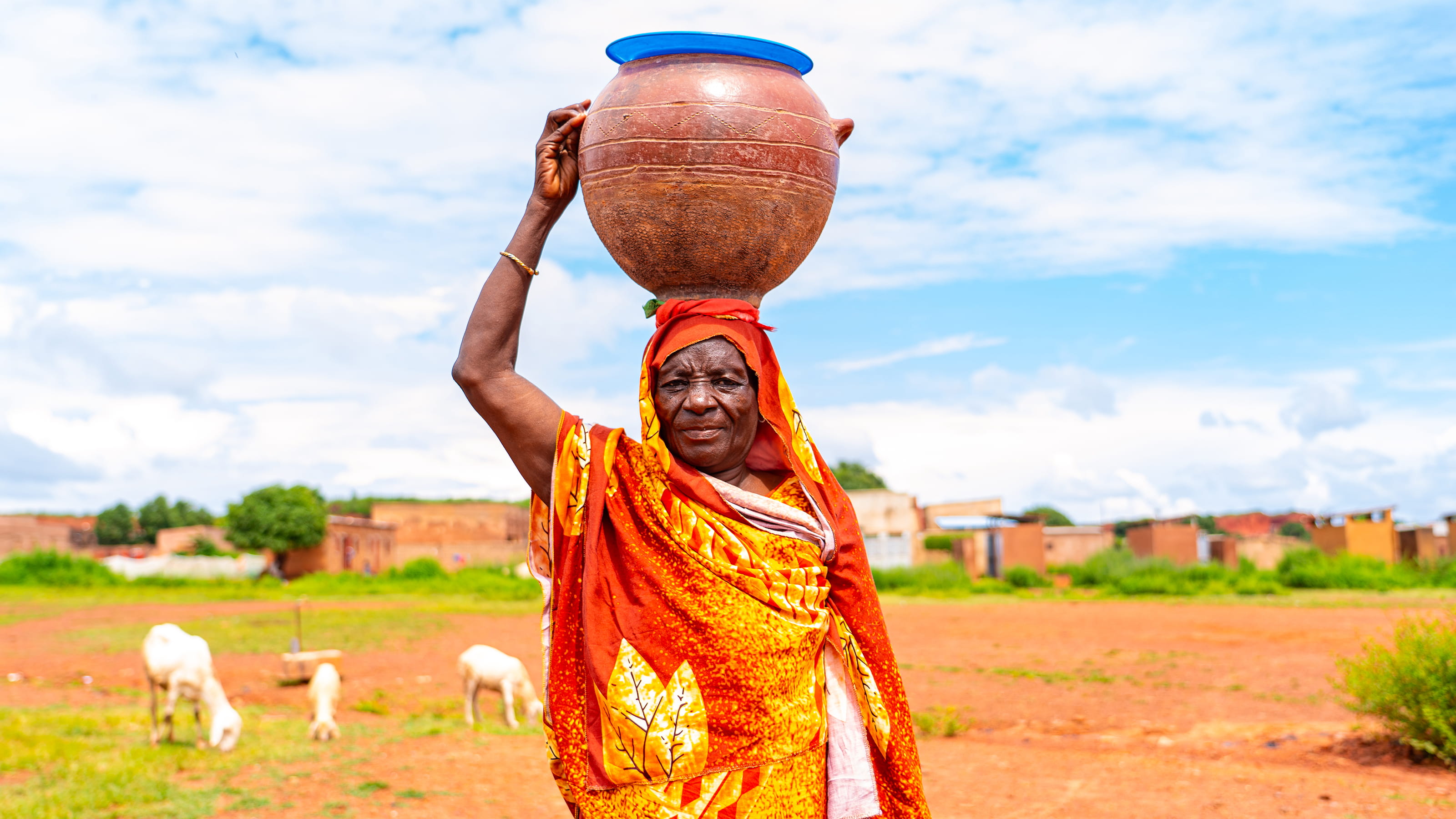 Someone wearing a bright orange scarf around their head and body carries a large pot on their head with one hand. 