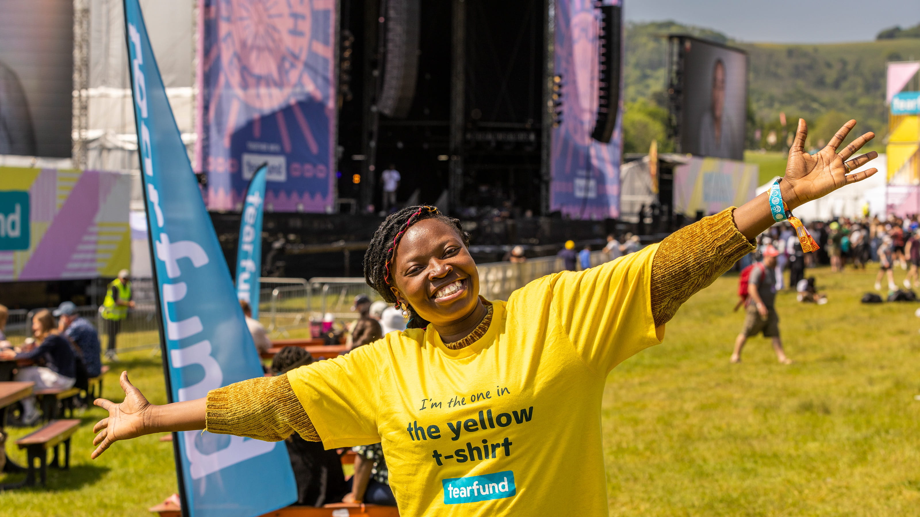 A smiling woman in a bright yellow Tearfund branded t-shirt in a field with a stage in the background