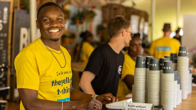 Smiling Black man in yellow shirt serving coffee with people in the background.