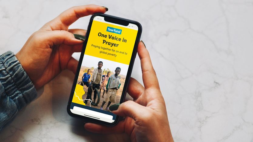 Hands holding a mobile phone with One Voice in Prayer displayed.