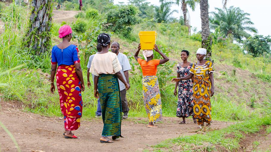 A group of women and one man walk on a dirt path in a rural, green area. One woman in the middle carries a yellow container on her head.