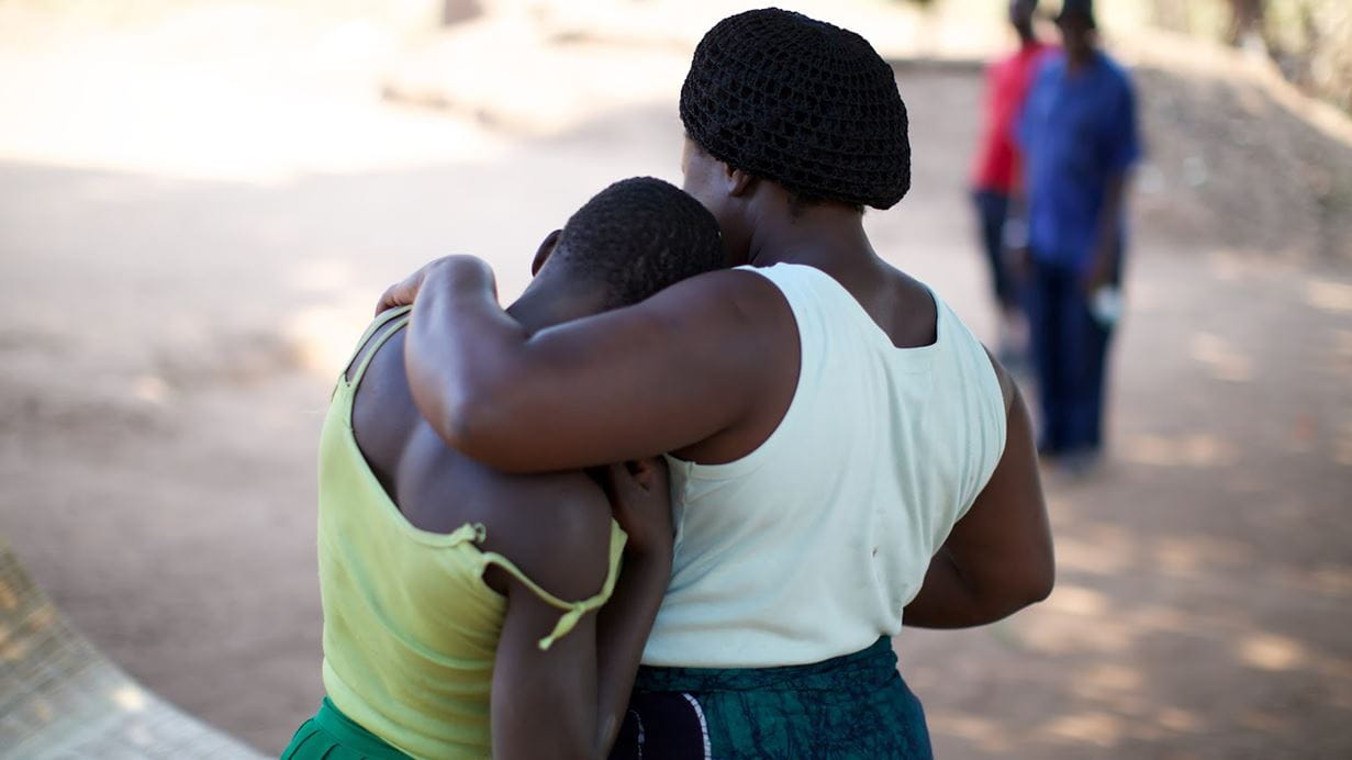 A Nigerian woman wearing a white top puts her arm around a girl wearing a light green strappy top, as two men in blue and red shirts look on in the distance. 