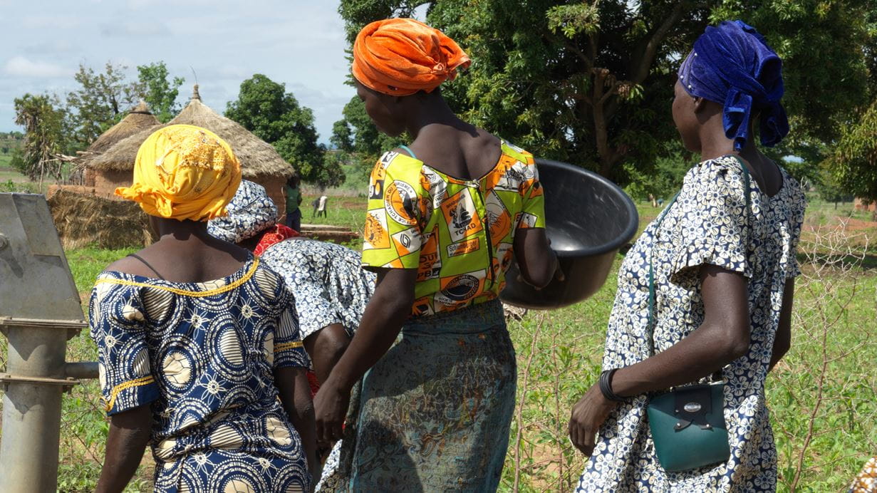 Three women from Chad dressed in colourful kitenge outfits queue to collect water from a pump in the village.