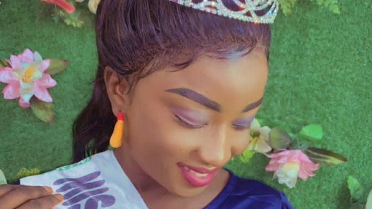 A woman from Malawi is wearing a small tiara, with delicate purple eyeshadow and pink lipstick. She is smiling warmly while looking down, wearing a navy blue top and yellow-orange drop earrings. The background features pink lotus flowers and green foliage. 