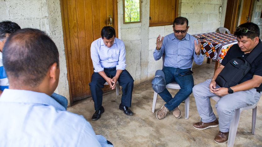 A group of men are on plastic chairs praying together outside, next to a building. One man lifts his hands up in prayer.