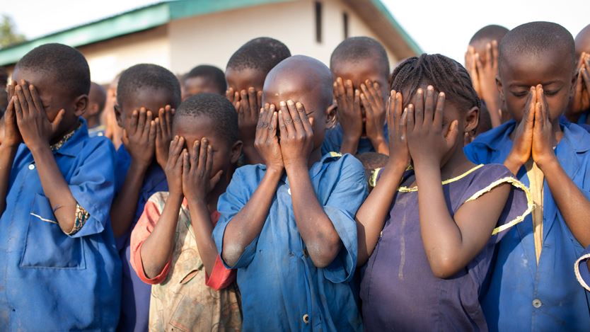 A group of children all hold up their hands to cover their eyes and pray.