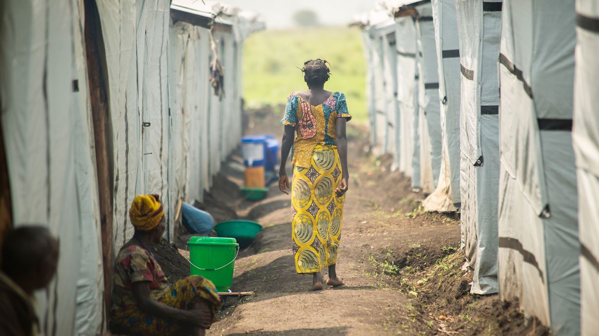 A woman wearing a patterned top and skirt walks with her back to the camera between rows of temporary shelters. Two people are sat on the ground next to shelters on the left.