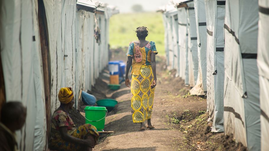 A woman wearing a patterned top and skirt walks with her back to the camera between rows of temporary shelters. Two people are sat on the ground next to shelters on the left.