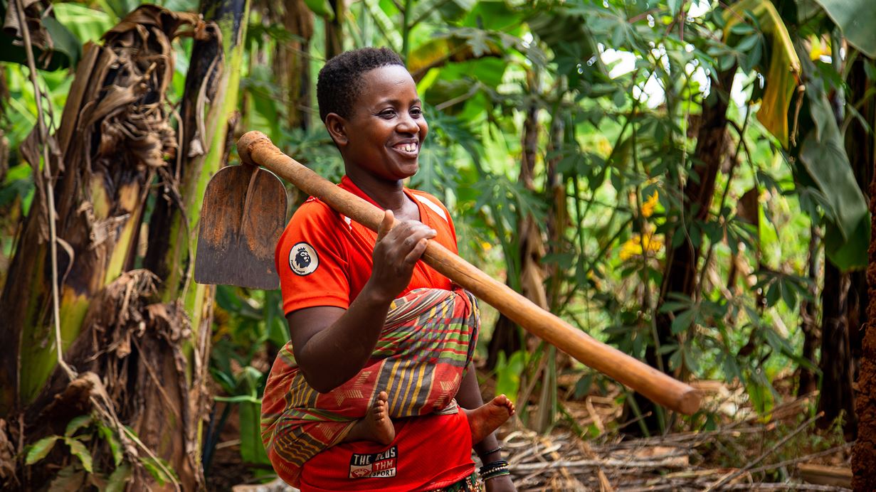 A woman with a young child wrapped in cloth on her back props a hoe over her shoulder and smiles broadly.
