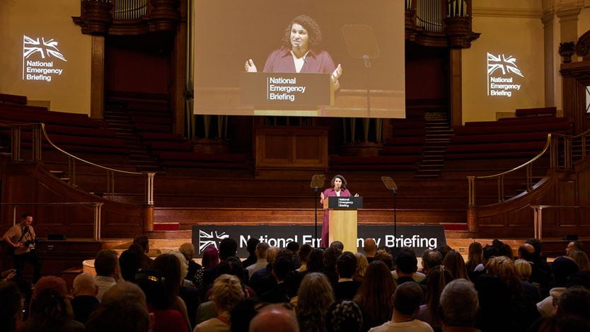 Woman speaks on a stage at the "National Emergency Briefing" to a crowd in auditorium, with a screen above.