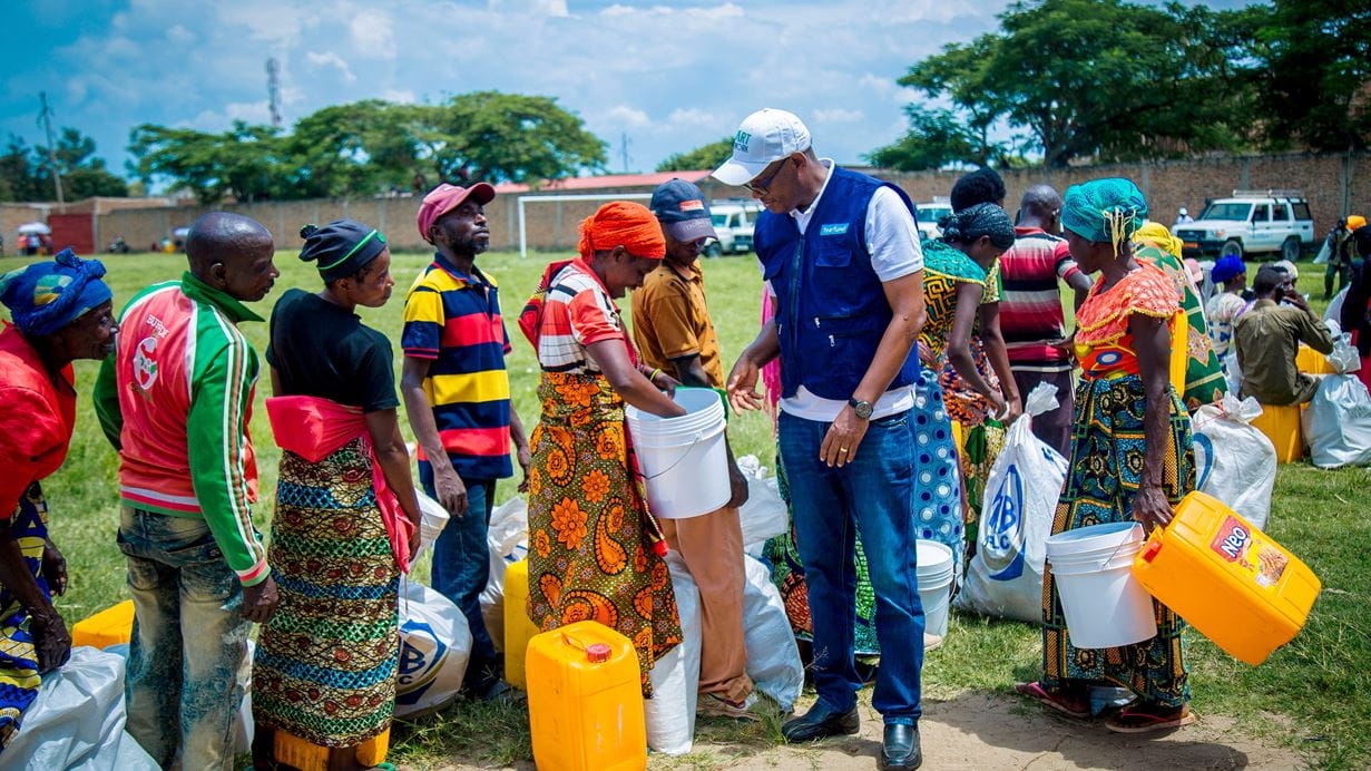 Burundian men and women holding buckets and jerrycans line up on a football field to receive vital aid supplies from a a Burundian Tearfund aid worker dressed in a white baseball cap, jeans and a navy-blue jacket.