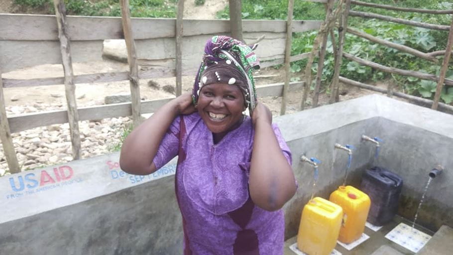 A woman smiling with water containers being filled from taps behind her