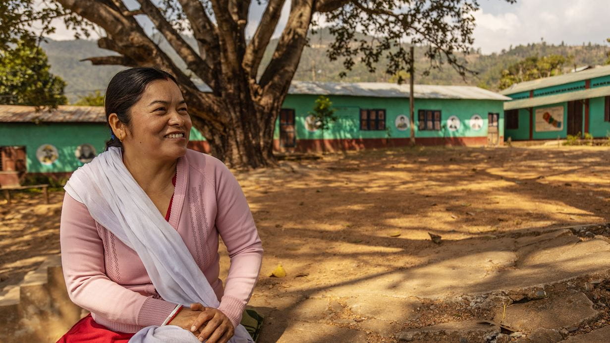 A woman sitting the shade of some trees with single story school buildings in the background