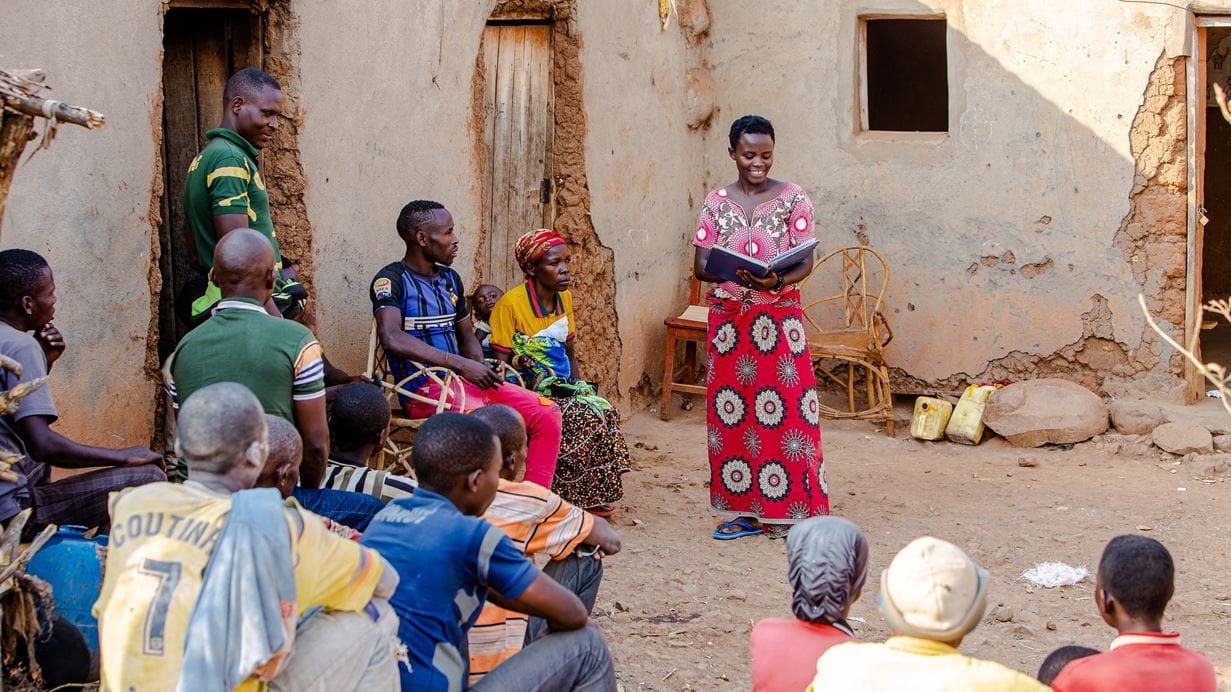 A group of people sitting down listening to a woman standing reading from a large book.