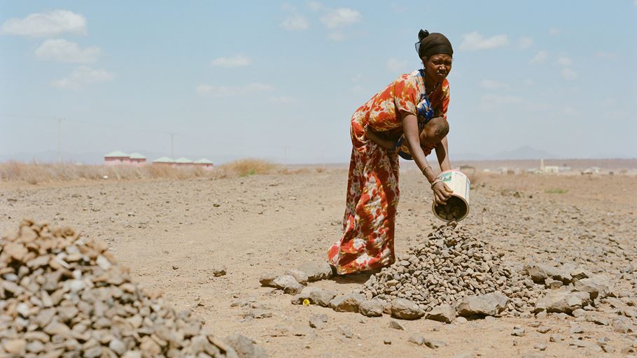 A woman with her child strapped to her chest, hammering rocks into ballast.