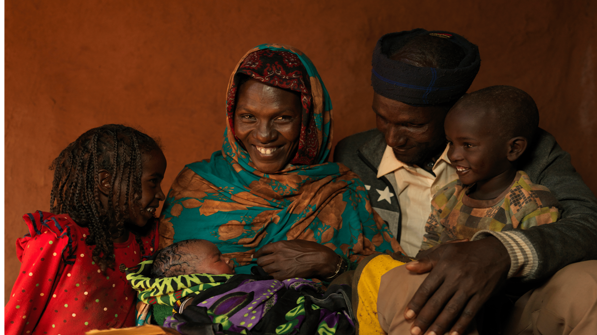 Godana holds one of his young sons while his wife smiles as she holds their newborn baby. One of their daughters also sits with them. They are in the shelter they have built from sticks, mud and a tarpaulin provided by the UN. The family had to leave their home after five years of drought in Ethiopia killed all their livestock and left them with no way to provide for themselves.