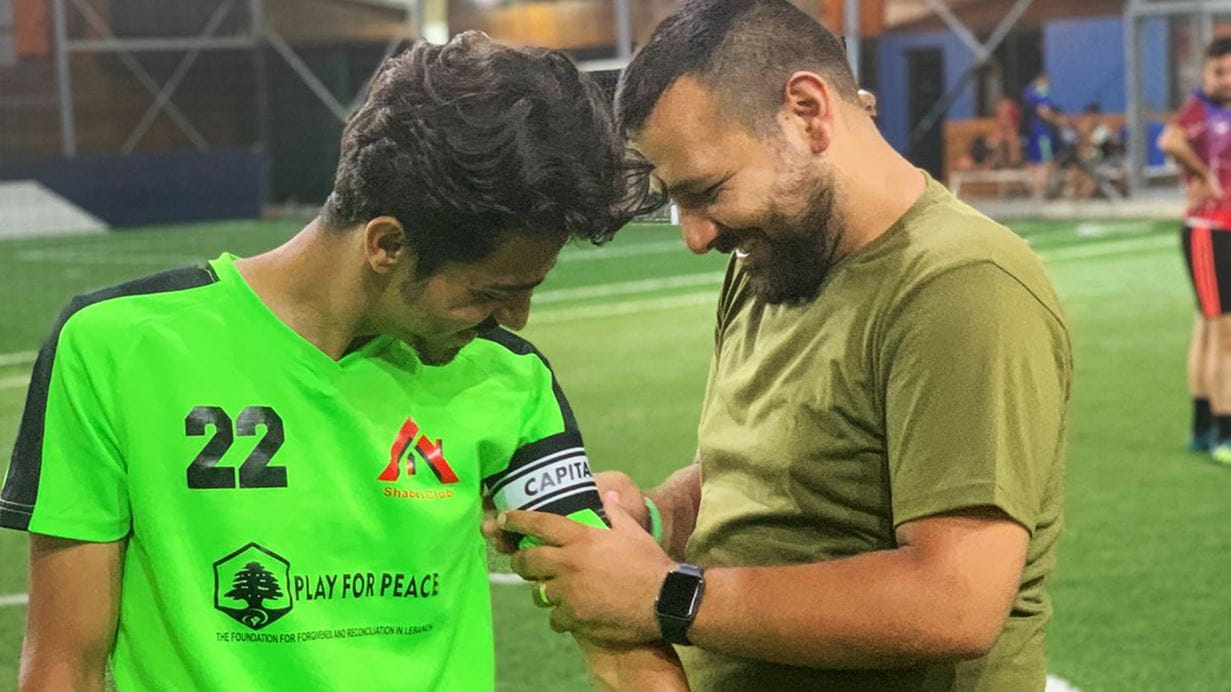 Emir, a Syrian refugee in Lebanon, wears football kit ready to play a match and smiles as his coach adjusts the armband showing that Emir is captaining the team.