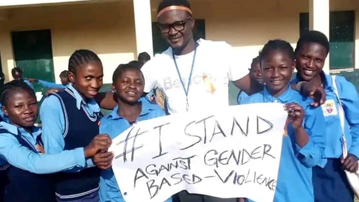 A group of people dressed in school uniform are stood holding a sign that reads '# I stand against gender based violence'