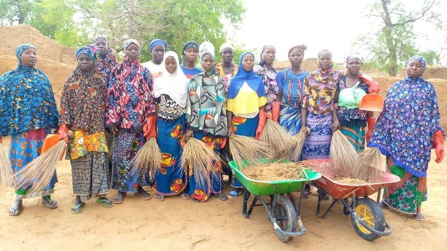 Mariam and her group of awareness-raising women stand together in the sunshine for a photograph with the wheelbarrows they use to clean up their community.