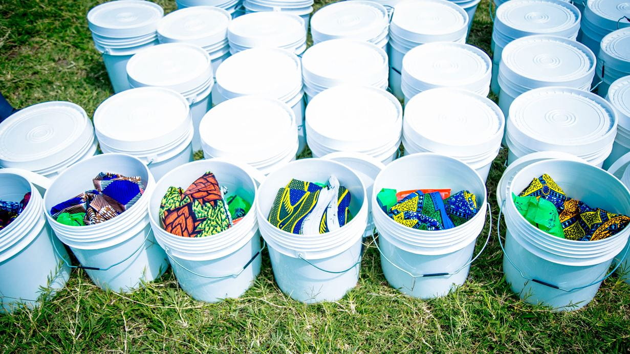 Rows of white buckets (useful for carrying clean water) stand ready to be distributed amongst people who have lost belongings because of the floods in Burundi. Inside the buckets are various essential items, including brightly-coloured blankets.