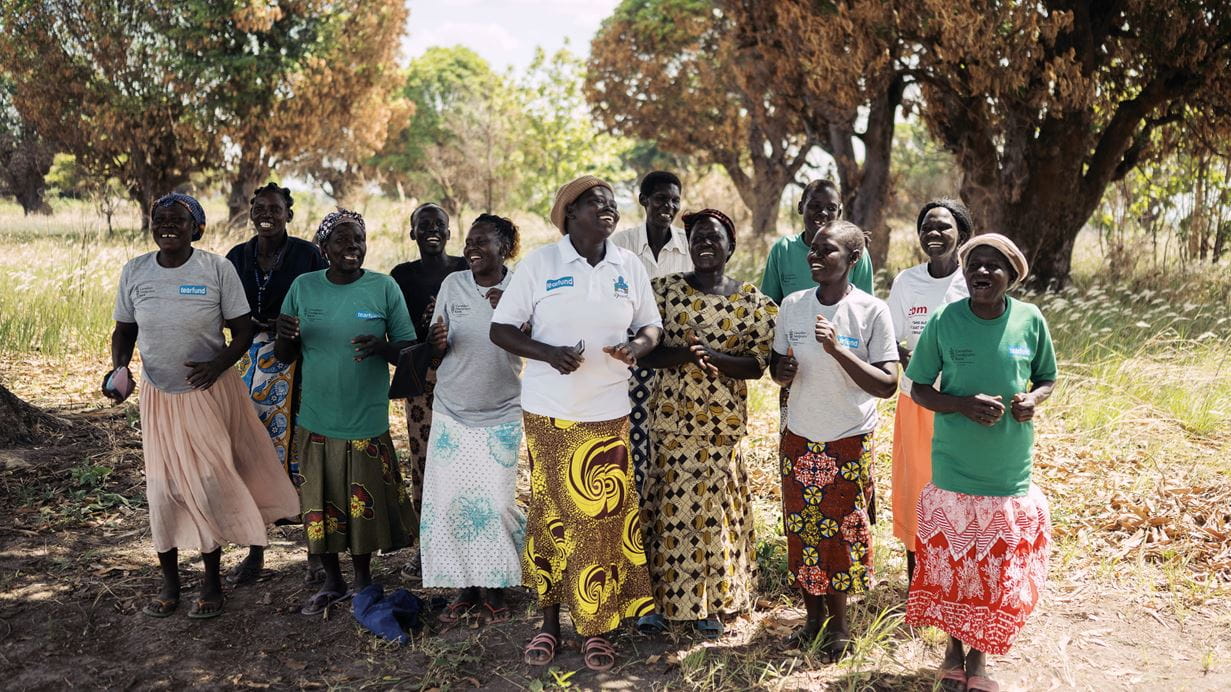 Mary and the members of her savings group stand outside in the shade of a large tree.