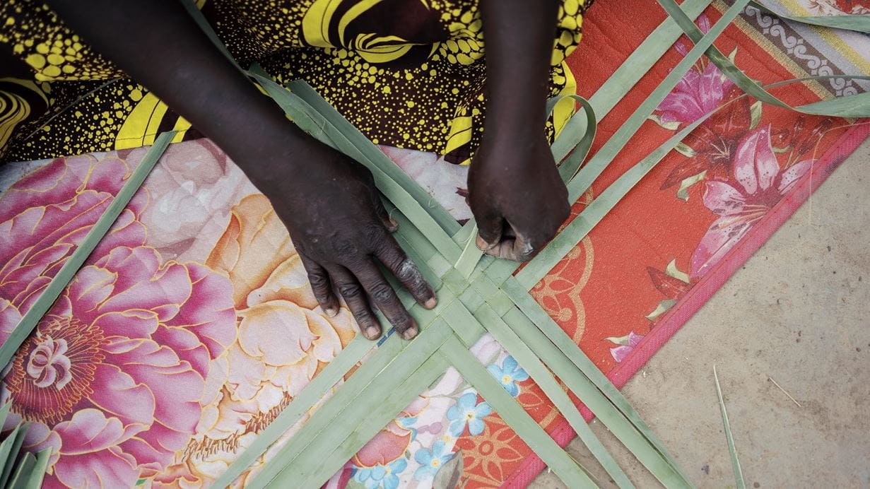 A close up picture of Mary's hands as she shows how she starts to weave a basket using pieces of flat grass. 
