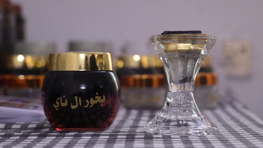 A small jar of incense with a gold lid and Maryam's logo in Arabic written on it stands on a checked cloth alongside a glass candlestick.