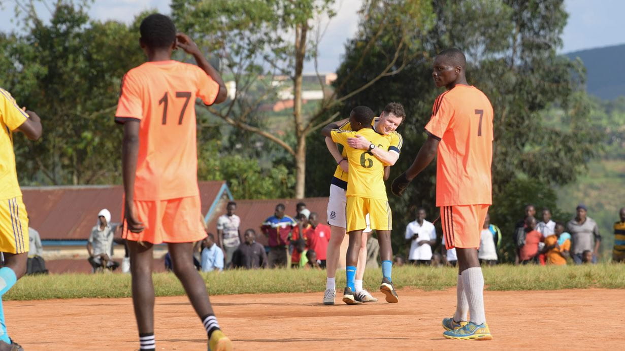 Several people on a football pitch dressed in football clothing. Two men embrace in the background.