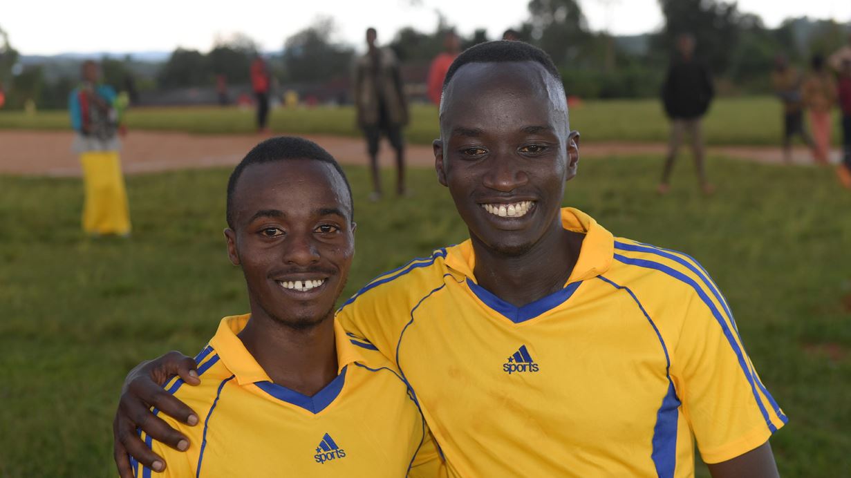 Two men in football tshirts put their arms over each other's shoulder and smile at the camera.