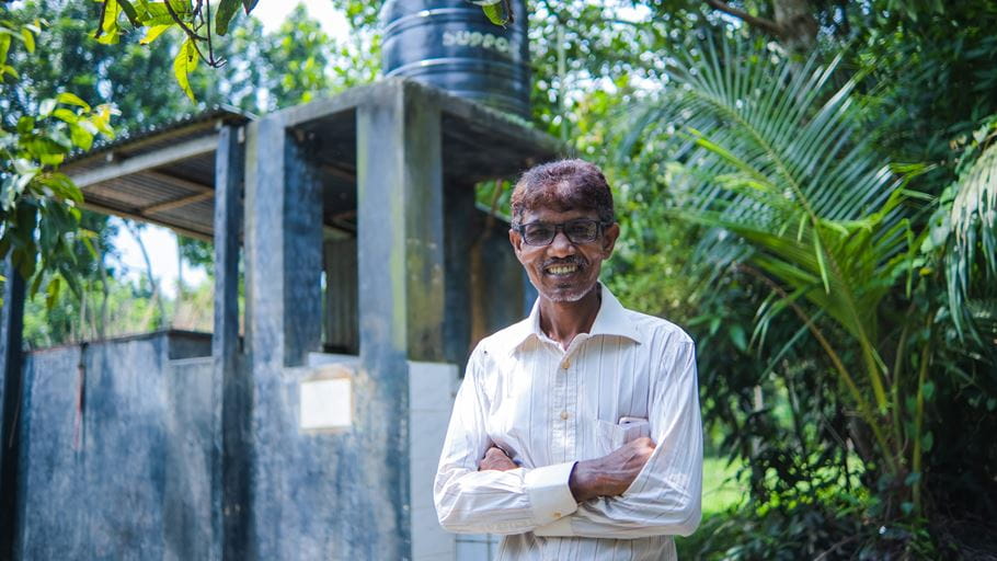 Pastor Sumon stands, smiling, in front of green trees and the water reserve built by his church with Tearfund support.