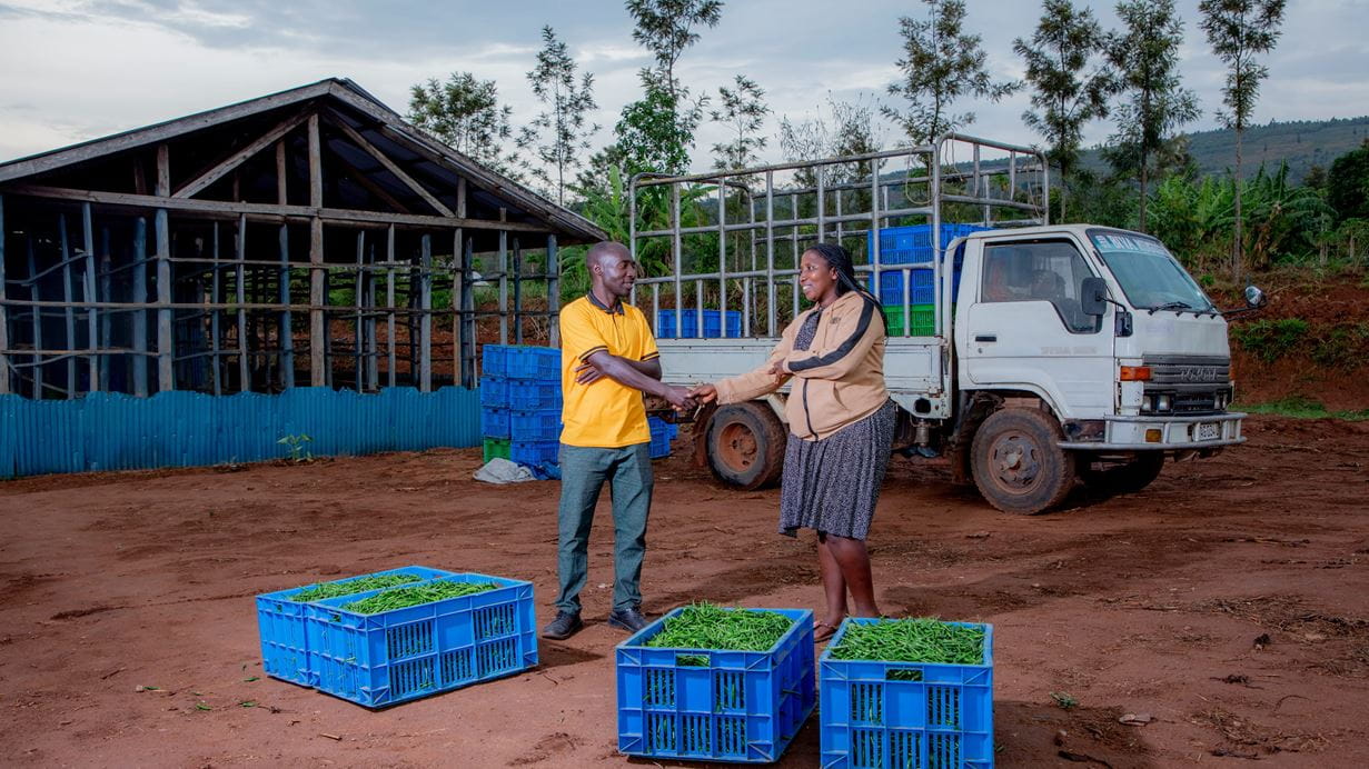 A man and woman shake hands by blue crates full of beans, standing in front of a white truck and a warehouse.
