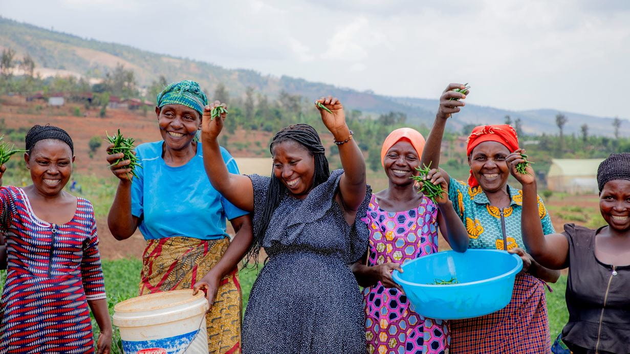 Six Rwandese women dressed in an array of colourful clothes laugh and smile as they proudly display their green beans amid the sloped Rwandan hillside.