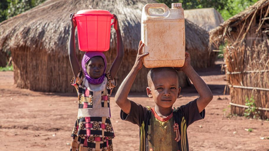 Ahamatou and Idrissa carry plastic containers of water balanced on their head.