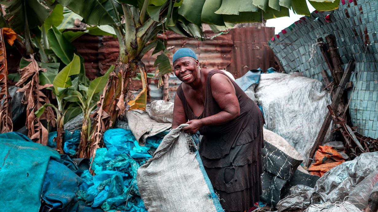 A woman stands outside next to bright green tree leaves. She holds a large sack containing plastic waste.